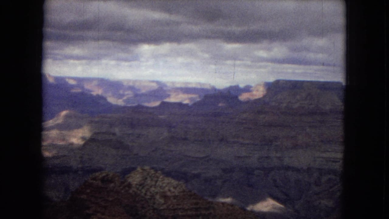 1967 Arizona home-movie frame with a wide view across the Grand Canyon, including dark ravines, distant plateau edges, and a cloudy sky with muted film color.