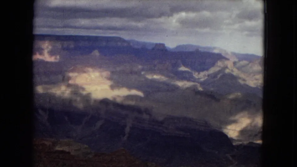 1967 Arizona 8mm still showing layered Grand Canyon walls and a broad overlook view, with sunlight breaking through clouds onto pale rock surfaces.