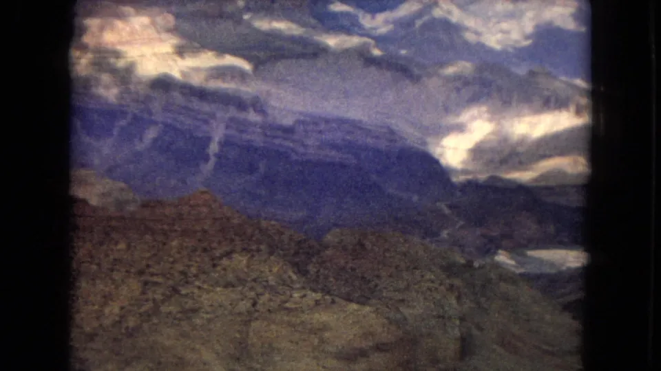 1967 Arizona home-movie frame showing rocky foreground terrain above a deep Grand Canyon view, with blue-gray canyon walls and bright cloud breaks overhead.