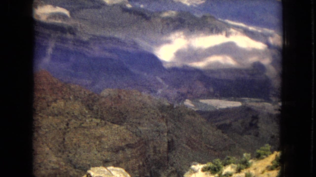 1967 8mm home-movie frame of a Grand Canyon overlook, showing layered canyon terrain, blue-toned distance, scattered clouds, and sunlit rocks in the foreground.