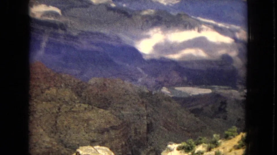 1967 8mm home-movie frame of a Grand Canyon overlook, showing layered canyon terrain, blue-toned distance, scattered clouds, and sunlit rocks in the foreground.