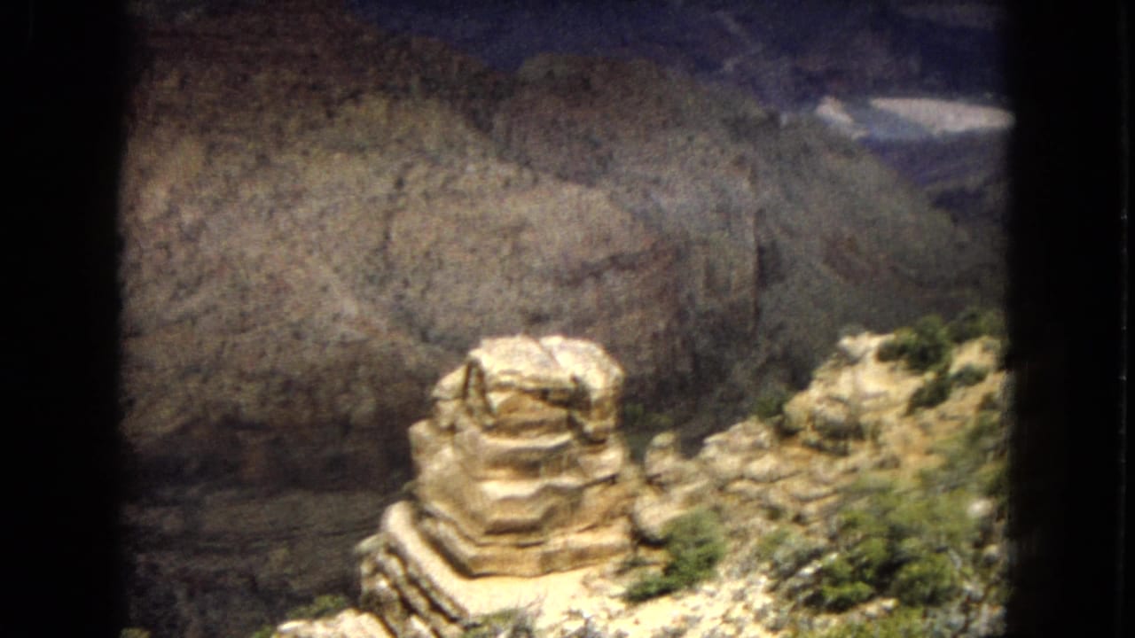 1967 8mm home-movie frame from the Grand Canyon area, with a prominent sunlit rock formation, scrub vegetation, and a steep canyon wall behind it.