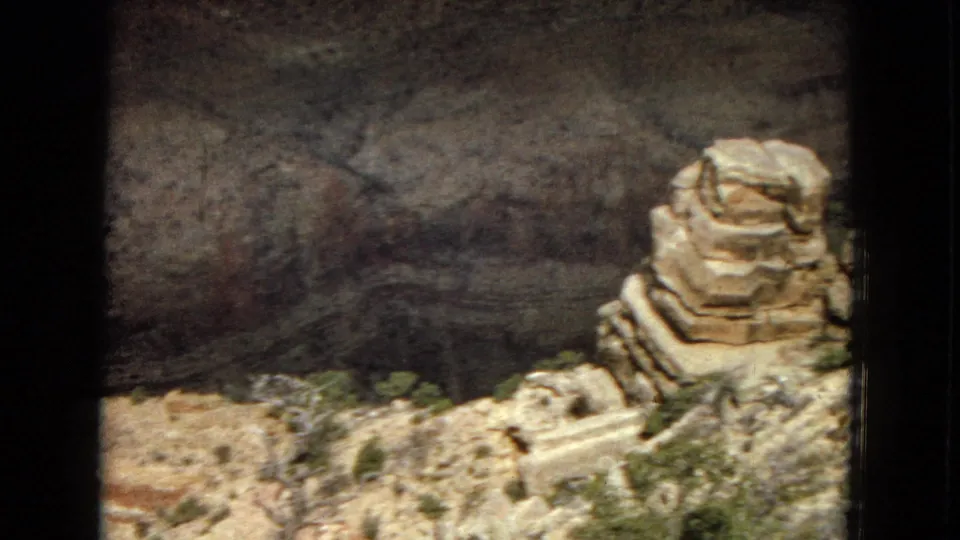 1967 8mm home-movie frame showing layered Grand Canyon rock ledges, a prominent foreground boulder, sparse desert plants, and a shadowed canyon drop beyond.