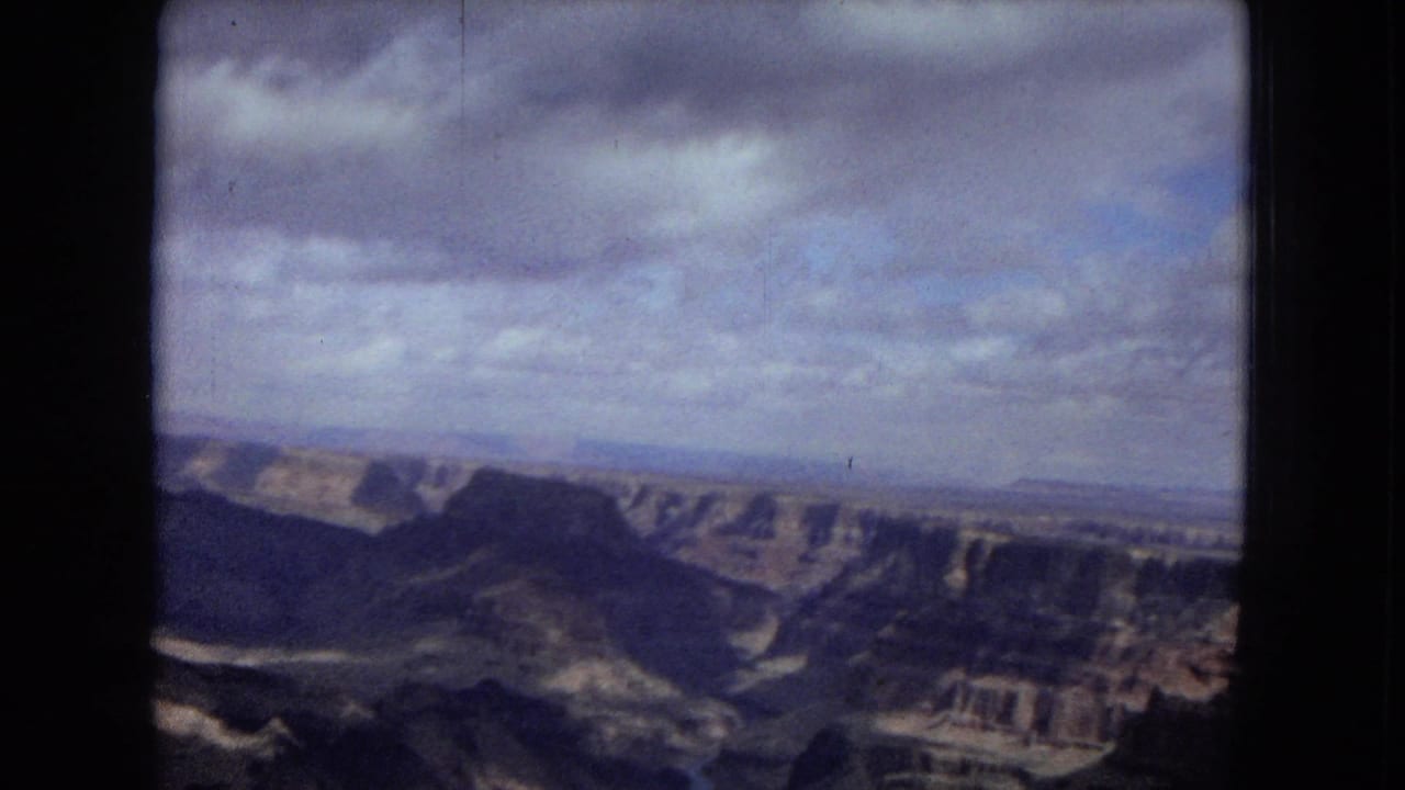 1967 8mm travel frame showing a wide canyon vista with layered cliffs, distant flatlands, cloud cover, and dark film borders.