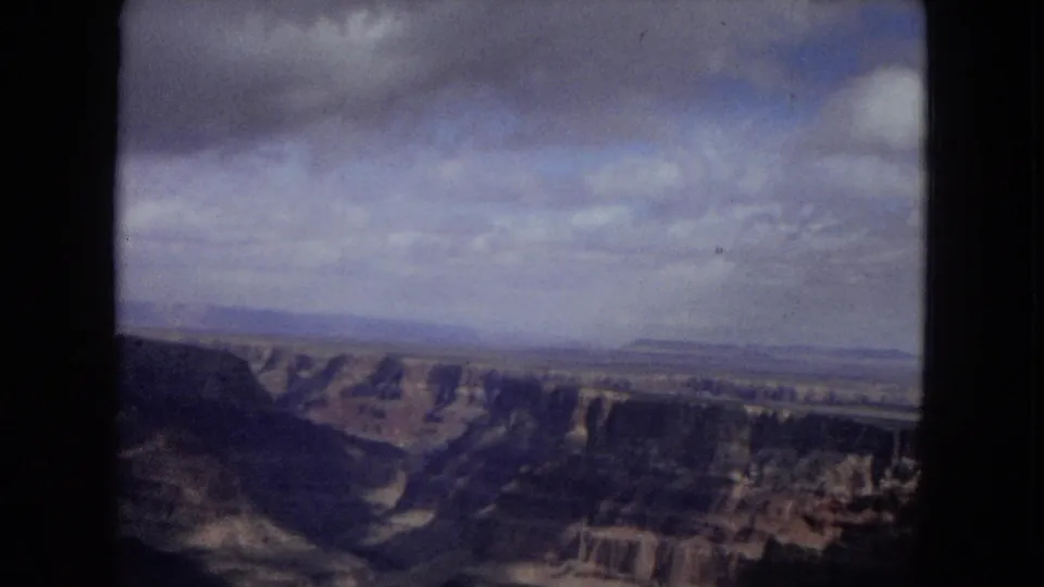 1967 8mm travel frame showing layered canyon walls, a hazy distant mesa line, and a partly cloudy sky from a visitor overlook.