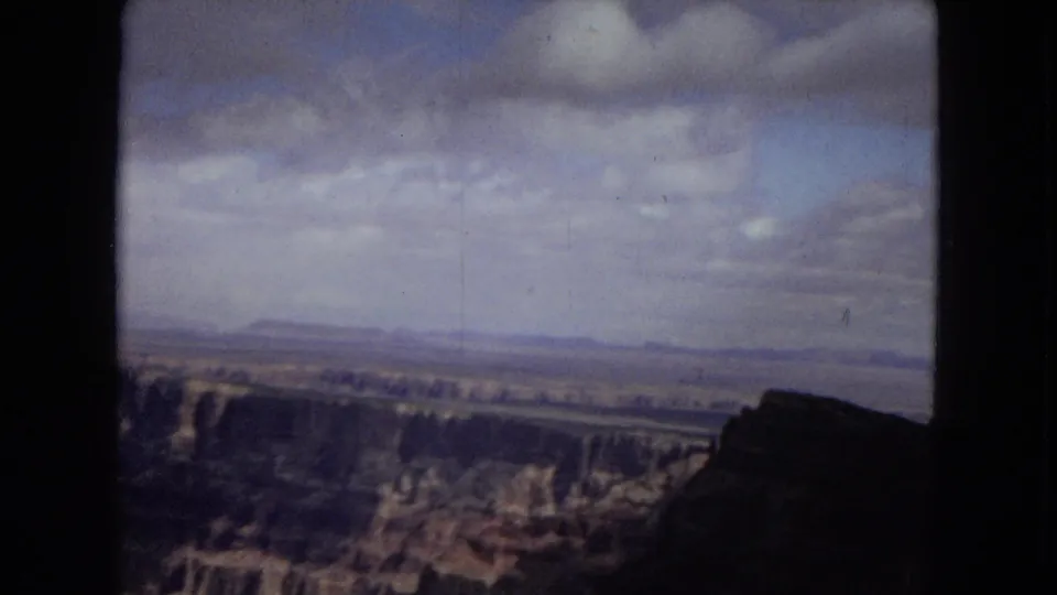1967 8mm travel frame showing a canyon rim, distant desert flatlands, cloudy sky, and a dark foreground rock silhouette.
