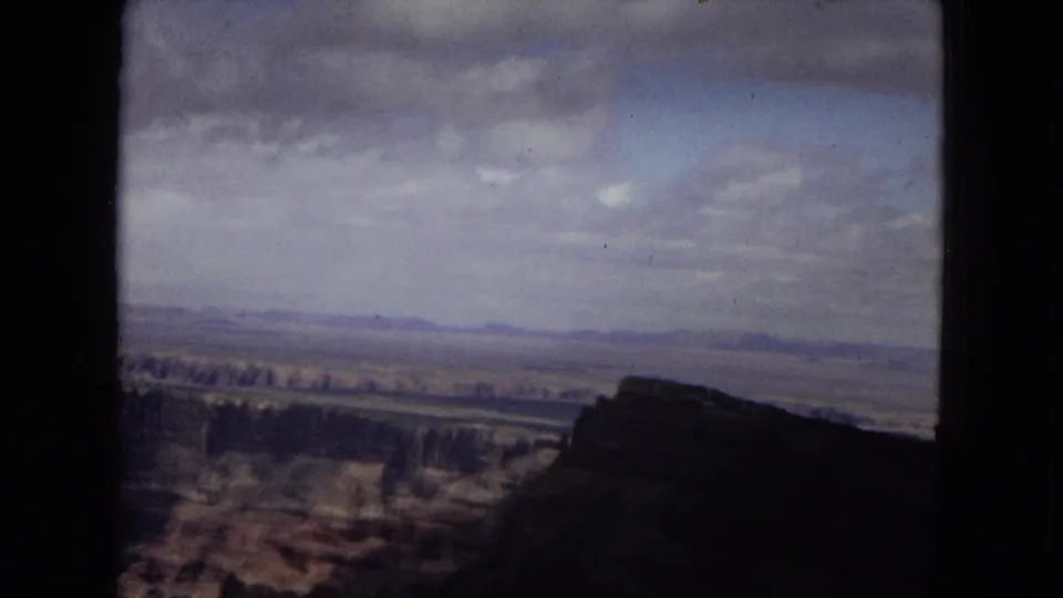 1967 Arizona 8mm view across the Grand Canyon, with a shadowed canyon edge in front and pale clouds over distant layered terrain.