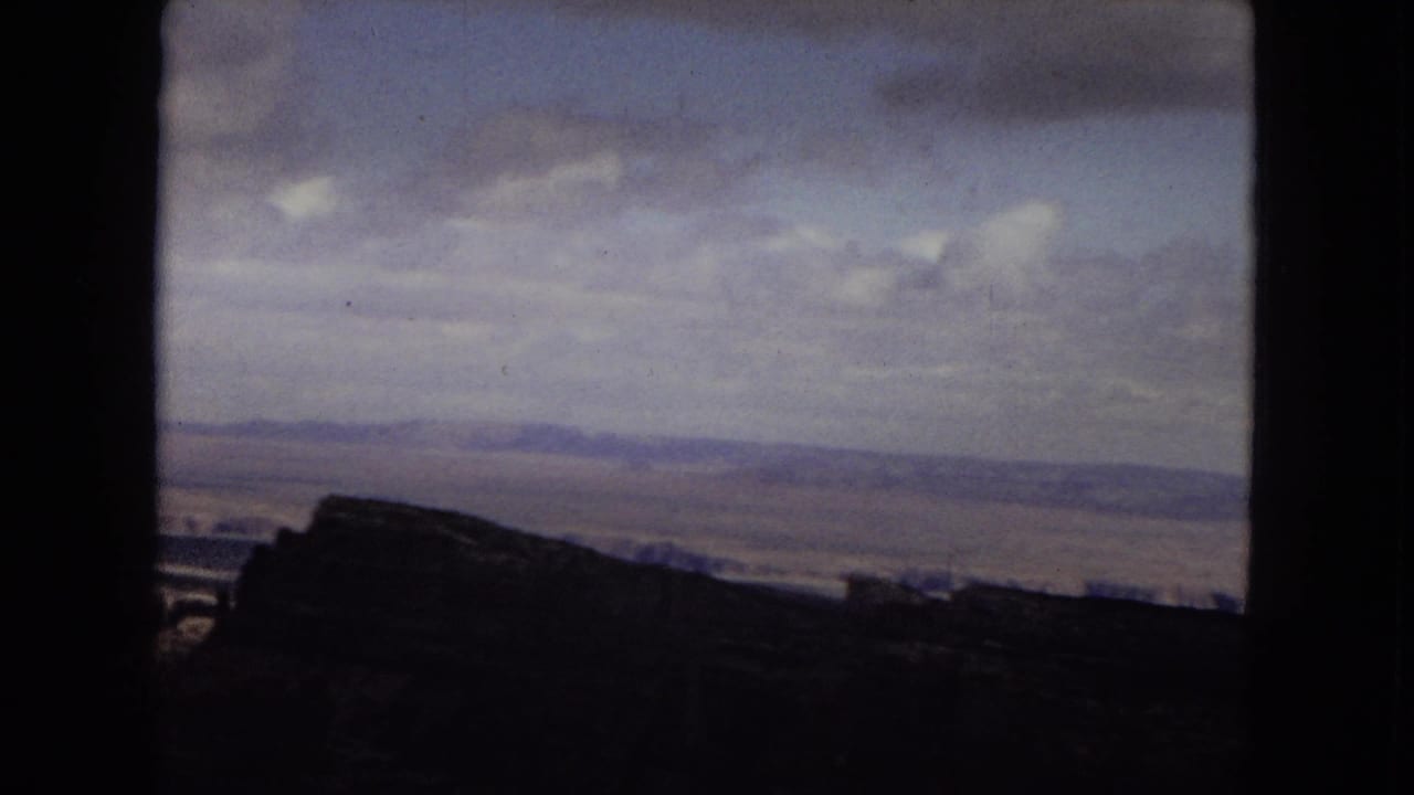 1967 Arizona home-movie frame looking across the Grand Canyon, with a dark rim silhouette and broad cloud cover over the distant plateau.