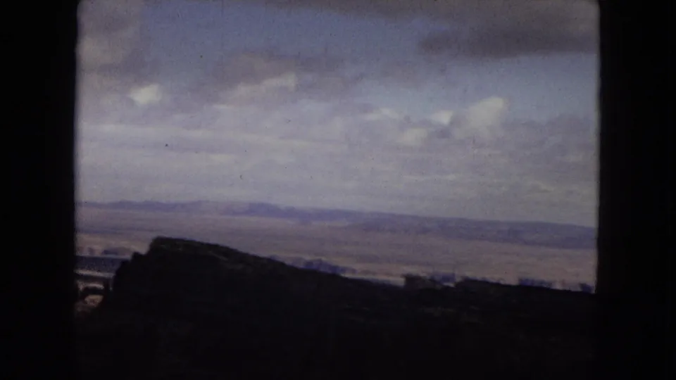 1967 Arizona home-movie frame looking across the Grand Canyon, with a dark rim silhouette and broad cloud cover over the distant plateau.