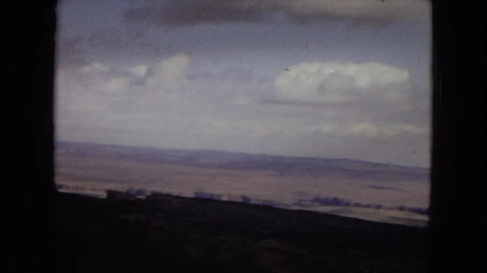 1967 Arizona 8mm landscape frame showing a low Grand Canyon horizon, dark foreground slope, and a large white cloud above the desert distance.