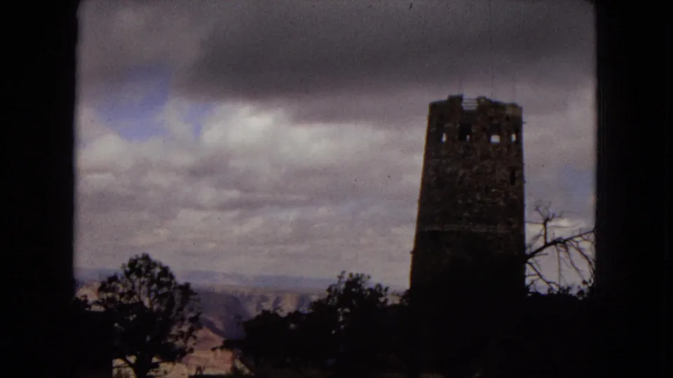 1967 United States travel footage showing a solitary stone observation tower near dark trees with distant hills and a cloudy sky behind it.