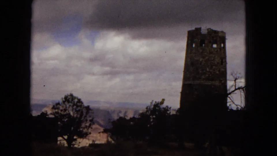 1967 Arizona 8mm travel frame showing the Desert View Watchtower at Grand Canyon National Park beyond silhouetted trees and a cloudy sky.