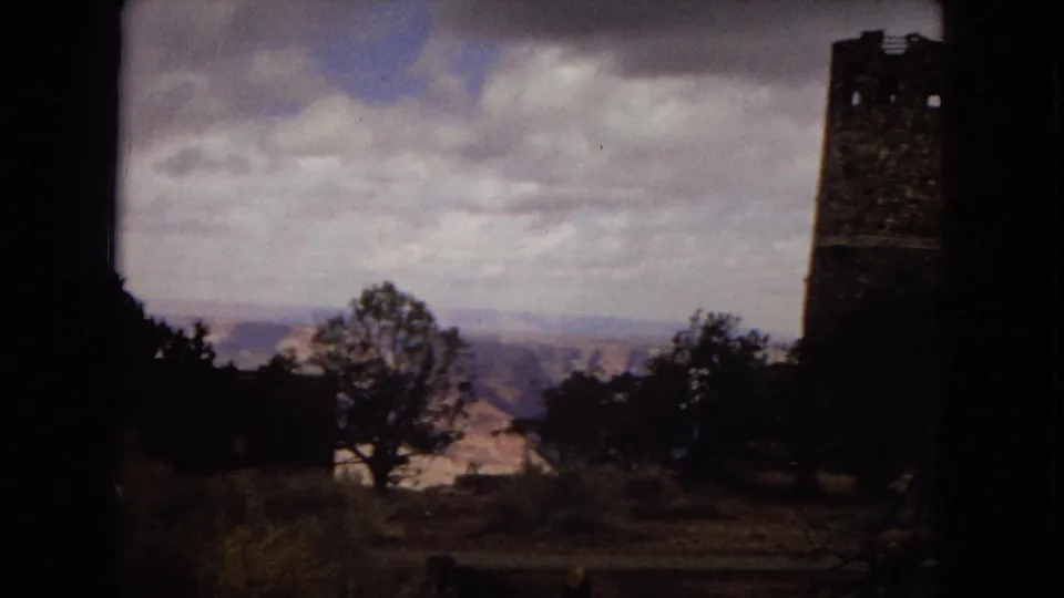 Grand Canyon-area travel view with the Desert View Watchtower partly at frame right, trees in the foreground, and distant canyon ridges under a cloudy sky.