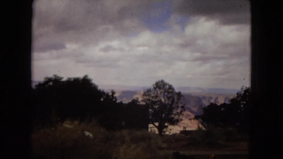 Arizona home-movie frame looking across trees and brush toward distant canyon terrain beneath a broad cloudy sky.