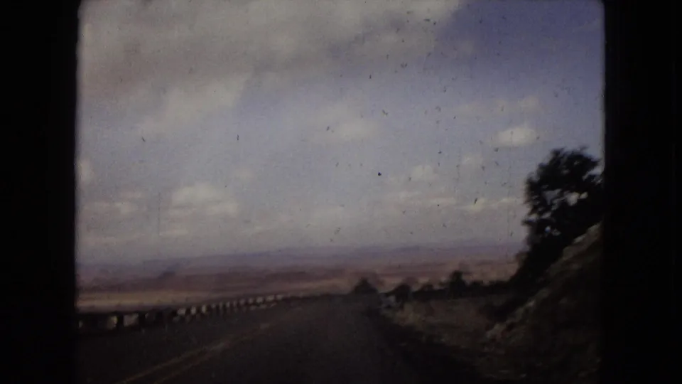 1967 Southwest road-trip frame from a moving vehicle showing a two-lane desert highway, roadside guardrail, open terrain, and hazy sky.