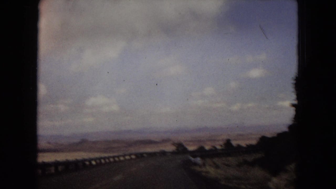 1967 home-movie view from a moving vehicle on a desert road with a guardrail, open arid landscape, and cloudy sky.