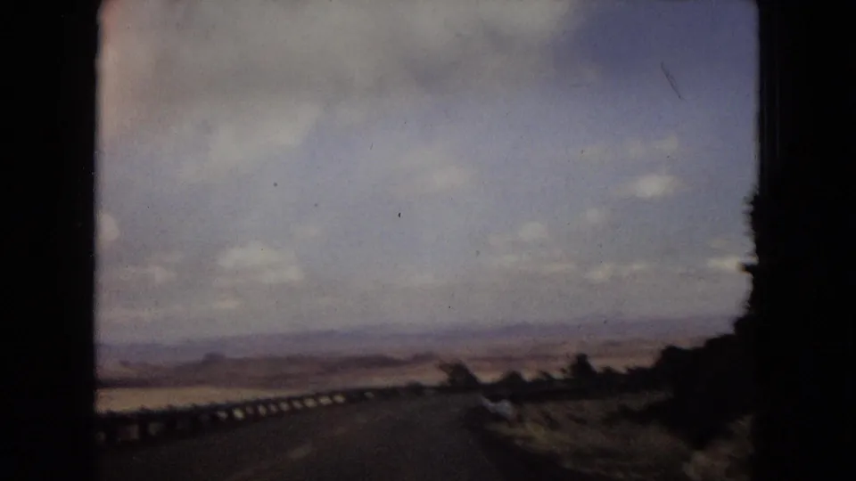 1967 home-movie view from a moving vehicle on a desert road with a guardrail, open arid landscape, and cloudy sky.