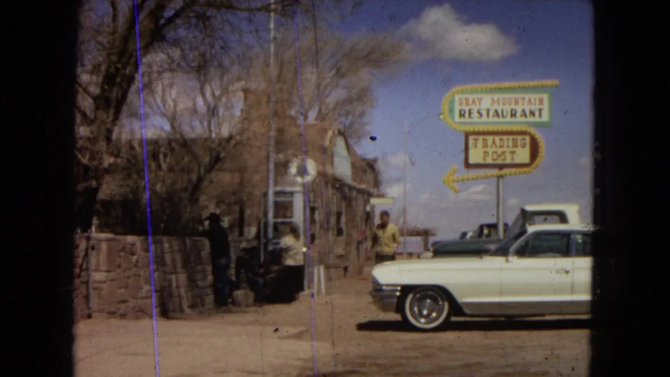 1967 roadside restaurant stop with people gathered near bags, a rustic stone building, a large Gray Mountain Restaurant and Trading Post sign, and a parked white car.
