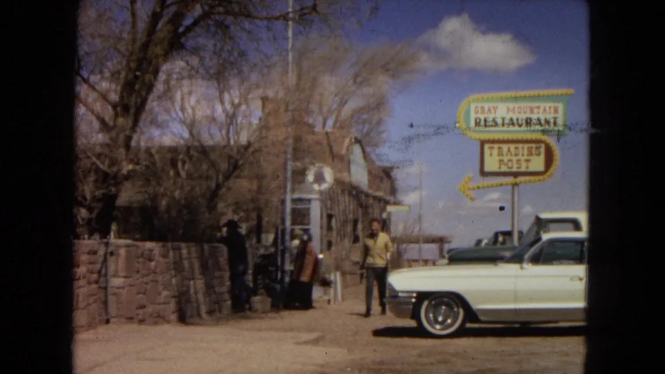 Roadside view of the Gray Mountain Restaurant and Trading Post sign with a person standing near a parked white classic car and others by the stone building.