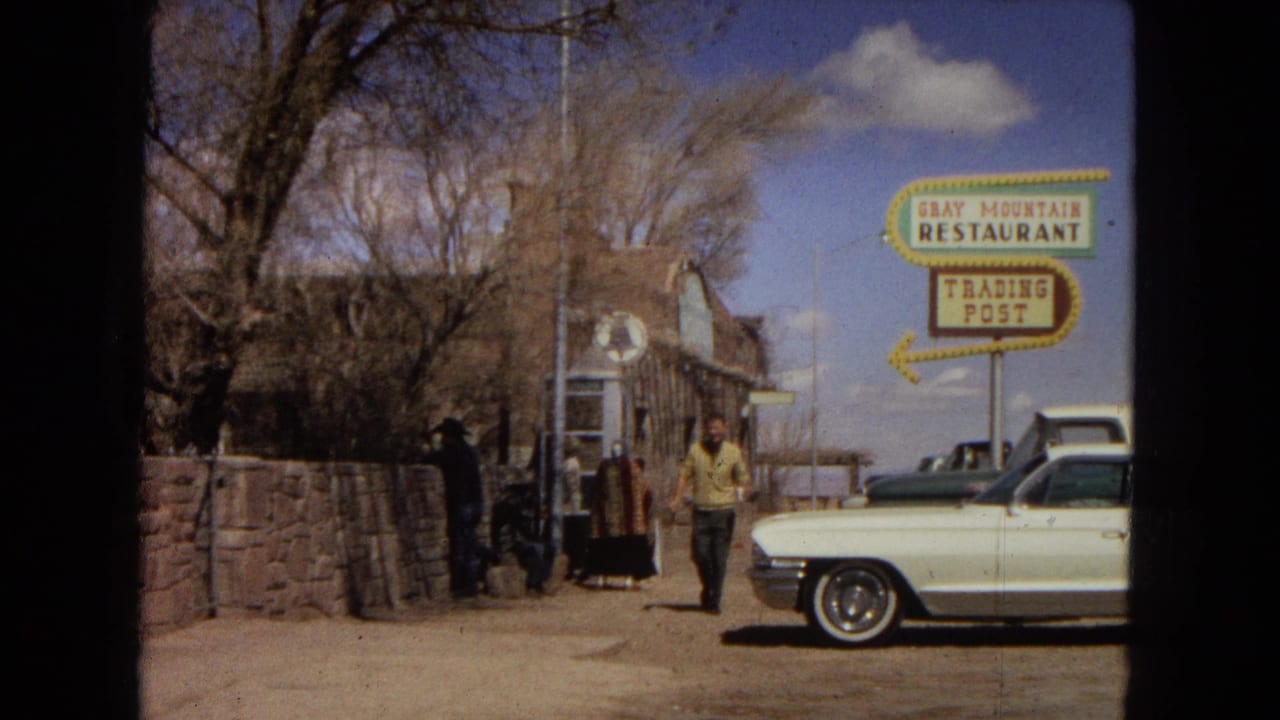 1967 Arizona home-movie frame showing a man in a yellow shirt walking near a white 1960s car beside the Gray Mountain Restaurant and Trading Post sign.