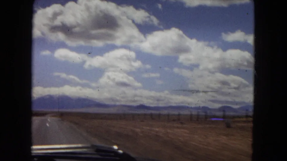 Point-of-view 8mm frame from inside a moving car on a straight desert road, with distant mountains and large clouds across the sky.