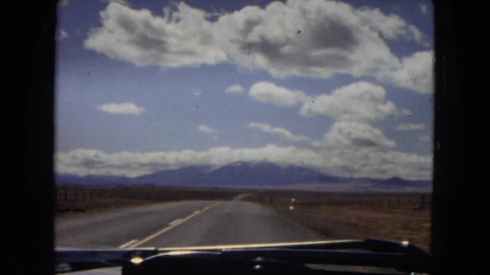 1967 Arizona travel footage frame showing an open highway from inside a car, with a broad desert plain, distant mountains, and dramatic cloud cover.