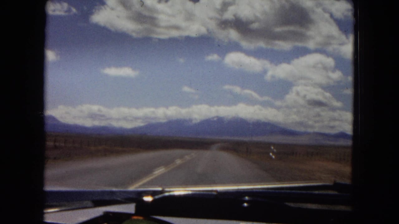 1967 Arizona home-movie frame showing a straight two-lane rural highway across open desert plains toward hazy blue mountains, with dashboard and windshield edges visible.