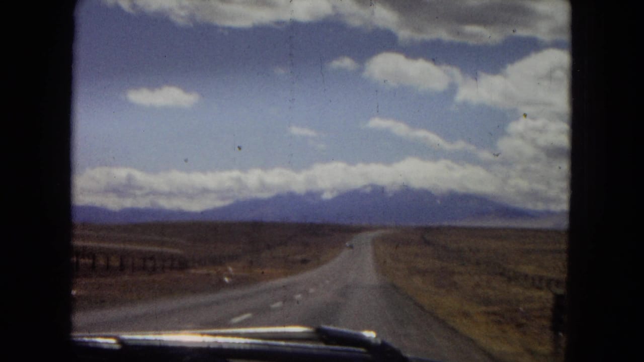 1967 Arizona road-trip view from inside a moving vehicle, with a paved highway running through dry open land toward cloud-covered mountains.