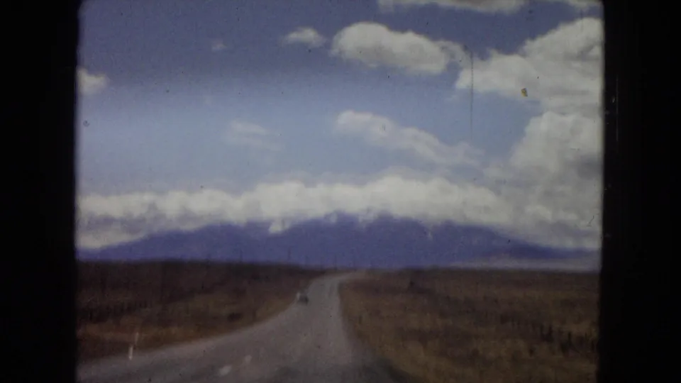 Windshield-shot 8mm frame from Arizona in 1967 showing a long rural road, dry roadside terrain, and a mountain range beneath large white clouds.