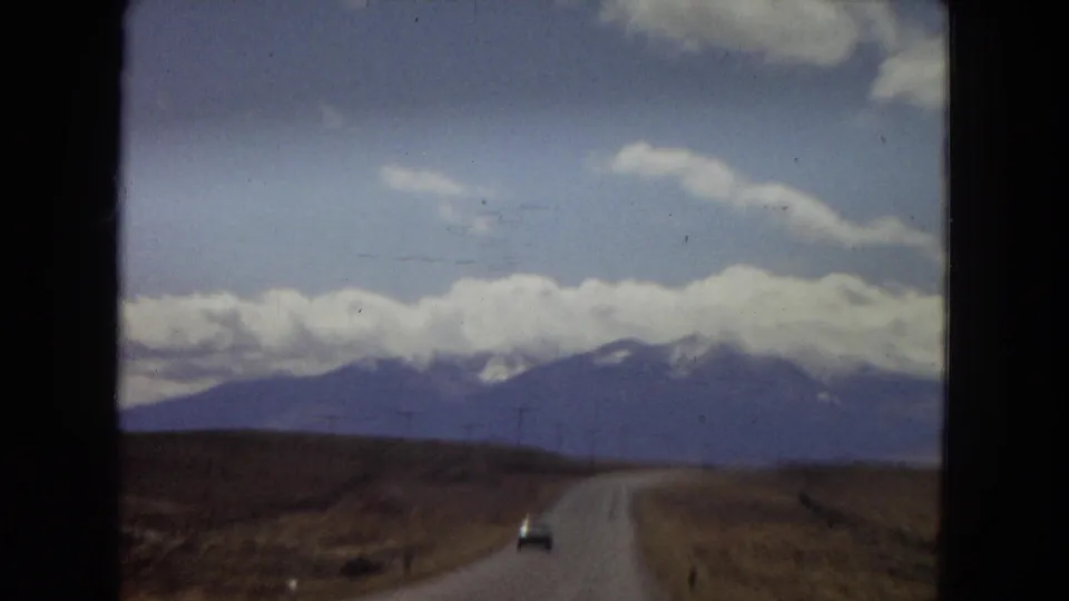 1967 Arizona home-movie frame showing a vehicle ahead on a two-lane rural road, with utility poles, dry fields, and blue mountains below a cloud-filled sky.