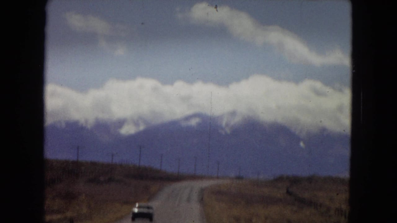 1967 Southwest road-trip view from a moving vehicle, with a curving road, small distant car, utility poles, and cloud-covered mountains ahead.