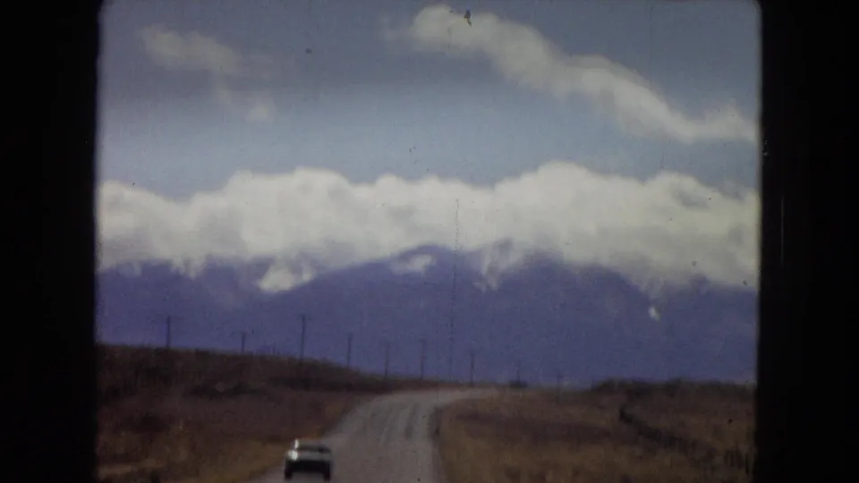 1967 Southwest road-trip view from a moving vehicle, with a curving road, small distant car, utility poles, and cloud-covered mountains ahead.