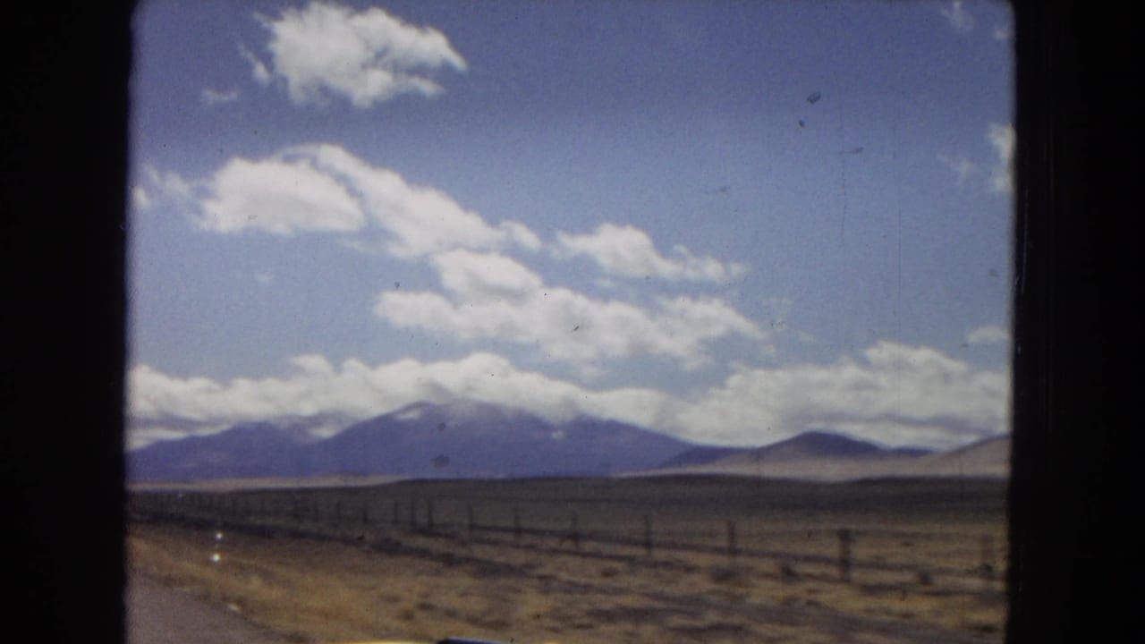 Arizona or Southwest home-movie view across dry open land with a roadside fence, low distant mountains, and scattered white clouds in a blue sky.