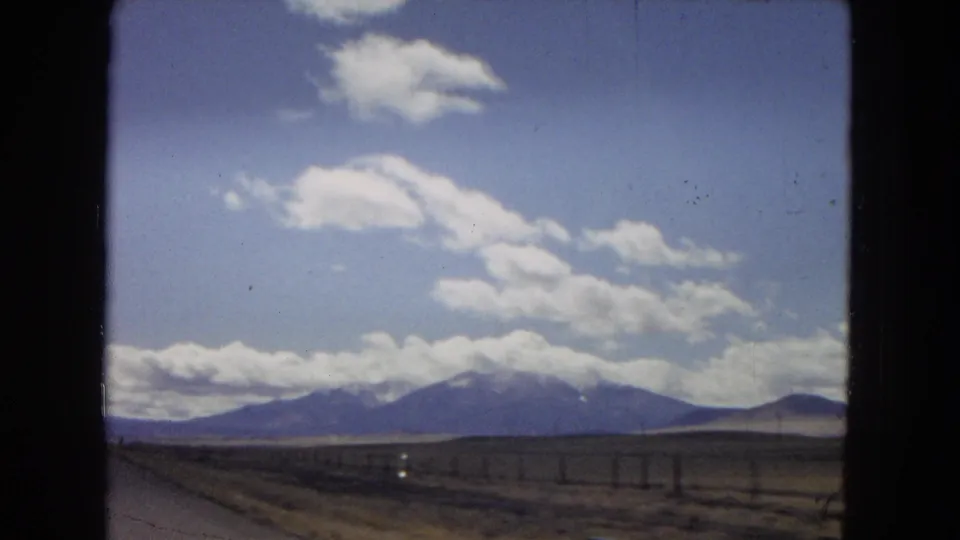 Moving-car view of a dry Southwest roadside, fence posts, distant mountain range, and bright cloud formations on faded 8mm color film.