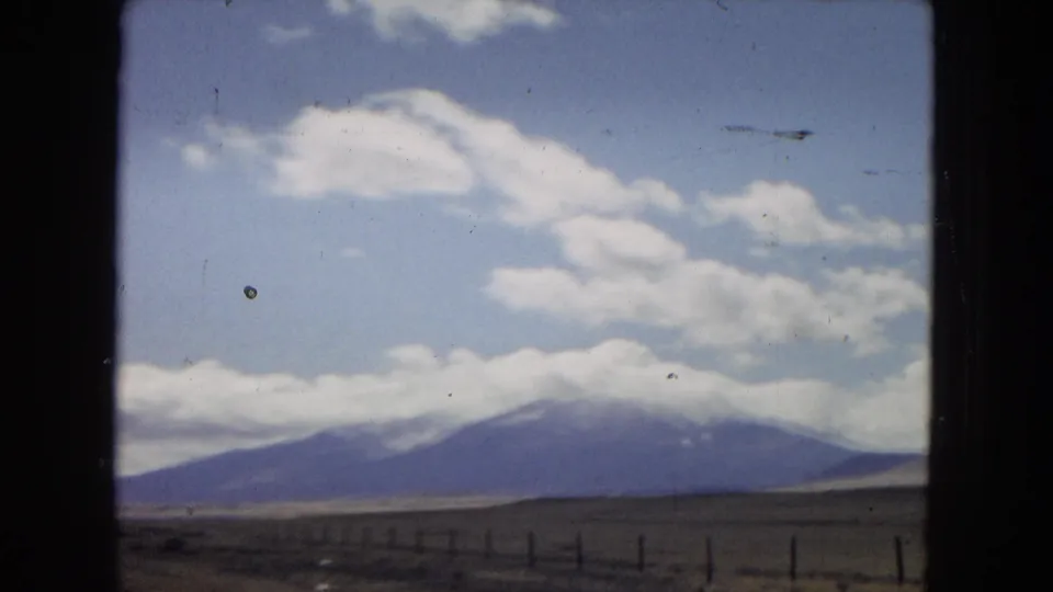 Wide 1967 road-trip landscape showing open desert foreground, roadside fence posts, a dark mountain range, and low white clouds across the peaks.