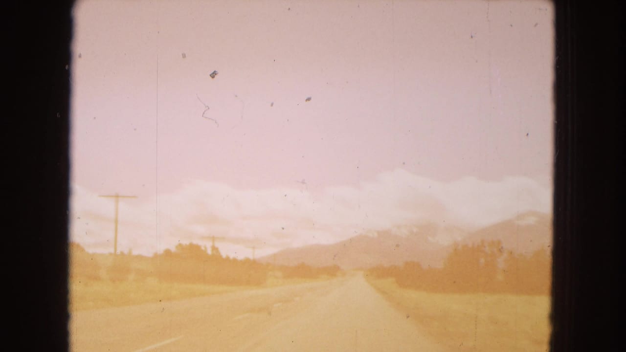 1967 Arizona 8mm frame showing a forward road view from a moving vehicle, with a two-lane highway, roadside utility poles, dry fields, and distant mountains under a pale pink sky.