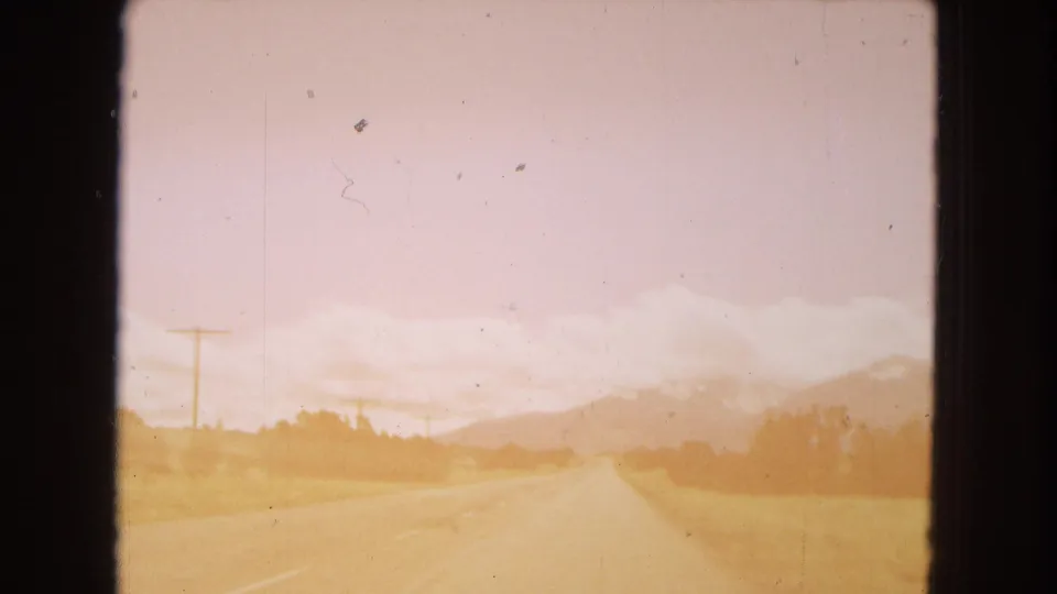 1967 Arizona 8mm frame showing a forward road view from a moving vehicle, with a two-lane highway, roadside utility poles, dry fields, and distant mountains under a pale pink sky.