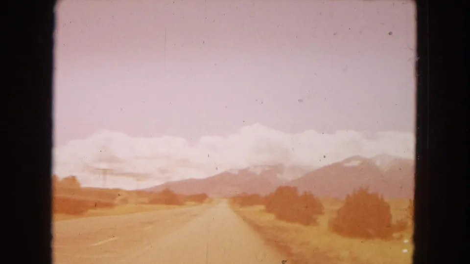 Warm-toned 1967 Arizona home-movie frame of a two-lane road, roadside shrubs, open yellow fields, and distant mountains seen through visible film-frame borders.