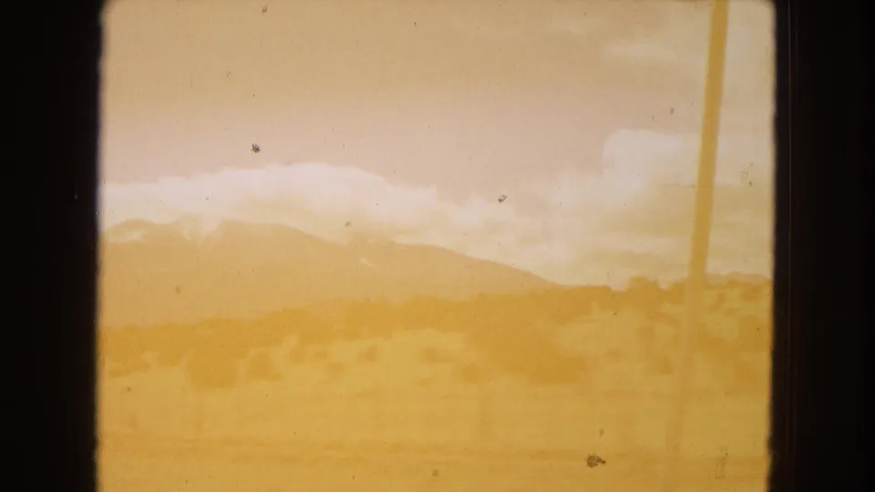 Soft, motion-blurred 8mm frame from the moving vehicle showing a golden roadside field, a utility pole at the edge, clouds, and distant mountains in Arizona.