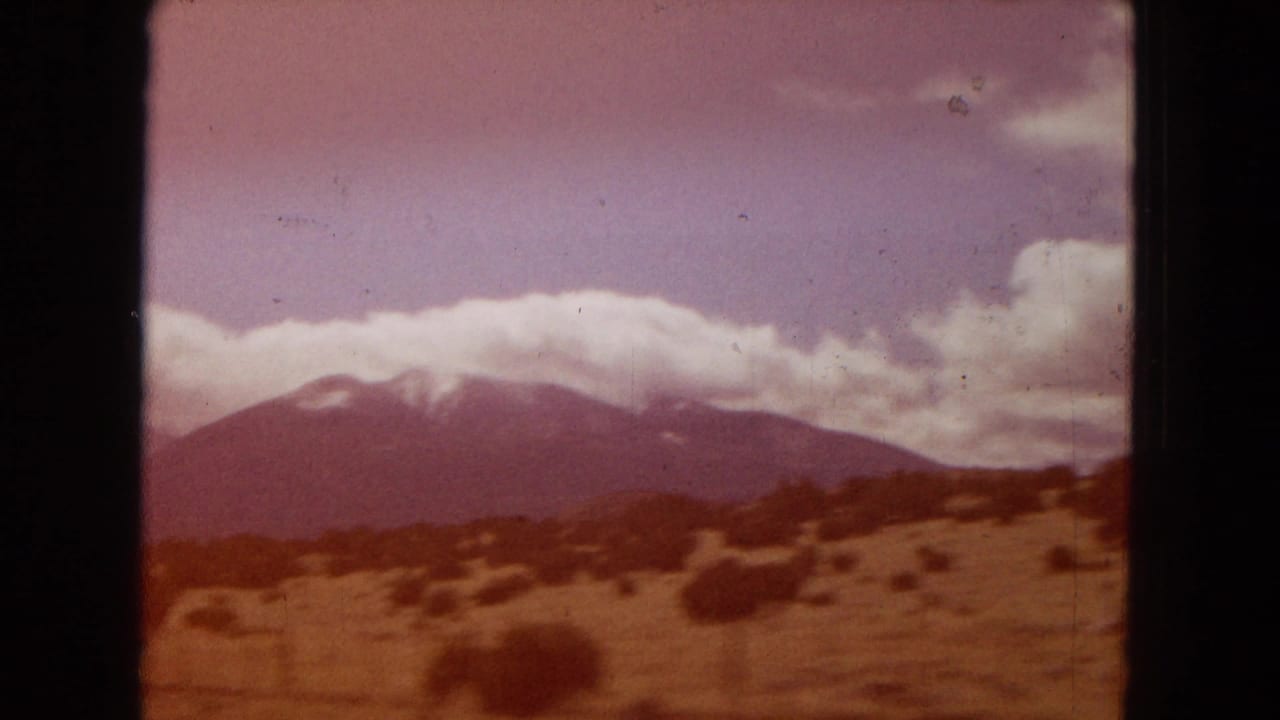 1967 Arizona home-movie frame showing a distant snow-dusted mountain range, dry open ground, scattered shrubs, and heavy clouds viewed through a dark vehicle or film edge.