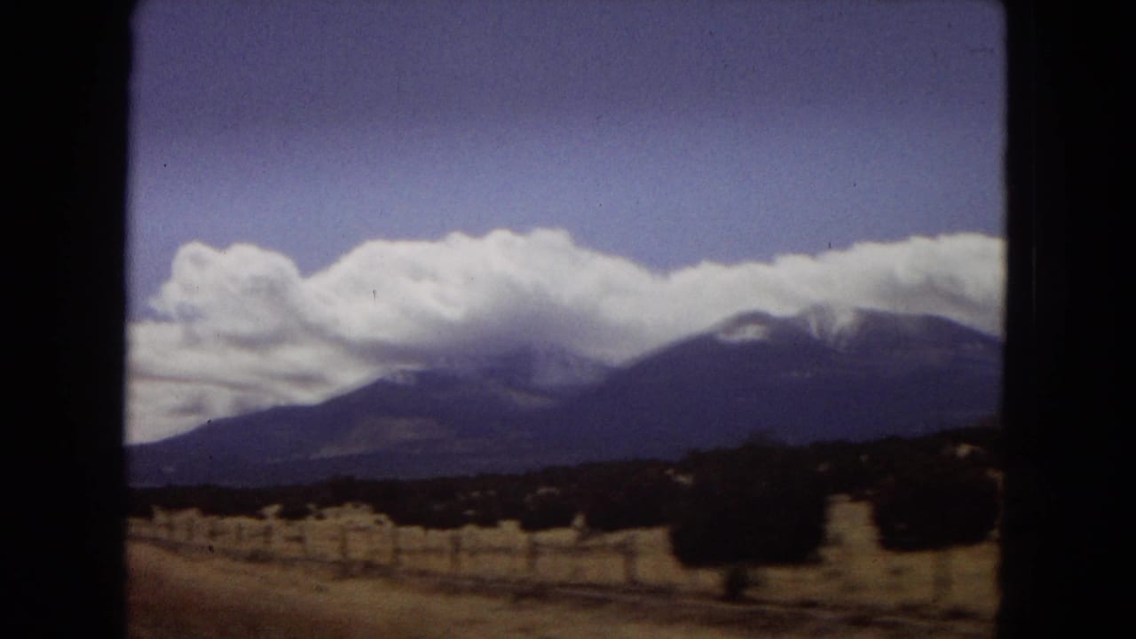 1967 Arizona travel footage frame with a dark window edge, roadside fencing or posts, dry vegetation, and a broad mountain range beneath bright white clouds.