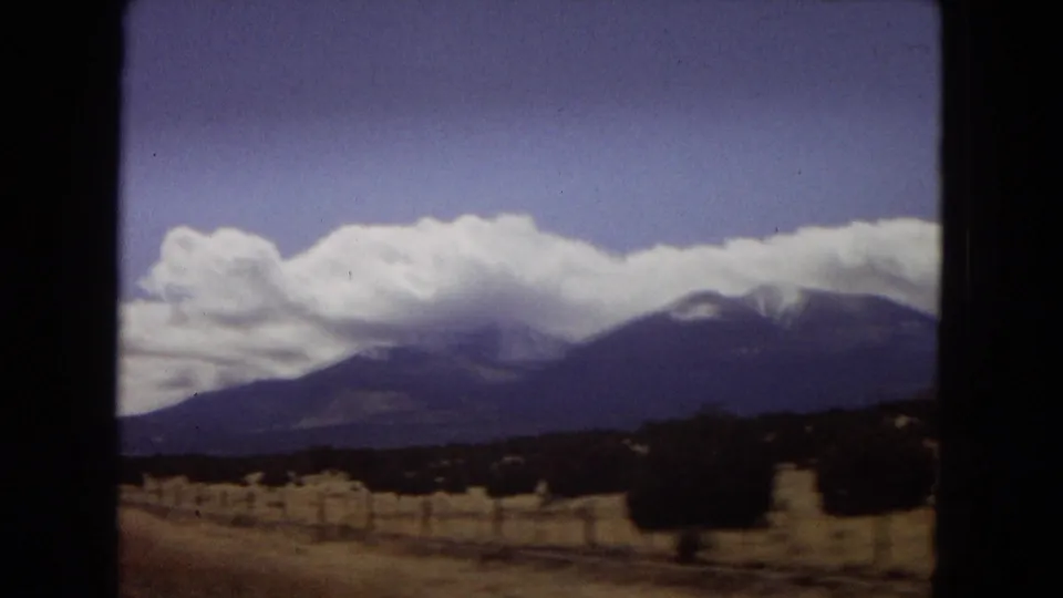 1967 Arizona travel footage frame with a dark window edge, roadside fencing or posts, dry vegetation, and a broad mountain range beneath bright white clouds.