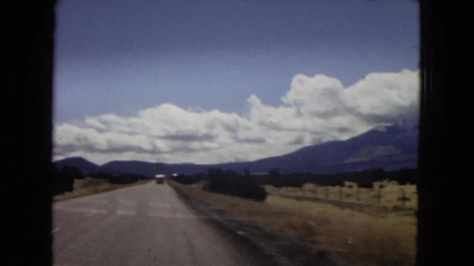 1967 Arizona home-movie frame looking down an open road toward distant mountains, with dry shoulders, low trees, and large cumulus clouds overhead.
