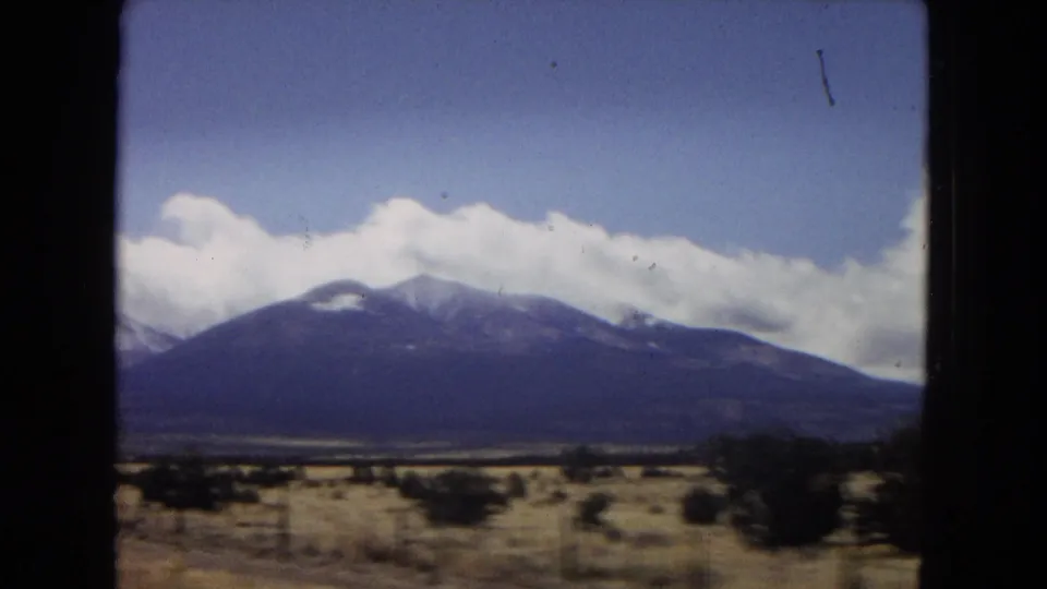 1967 Arizona landscape frame showing a broad snow-streaked mountain range beyond dry open ground, scattered shrubs, and bright clouds in a blue sky.