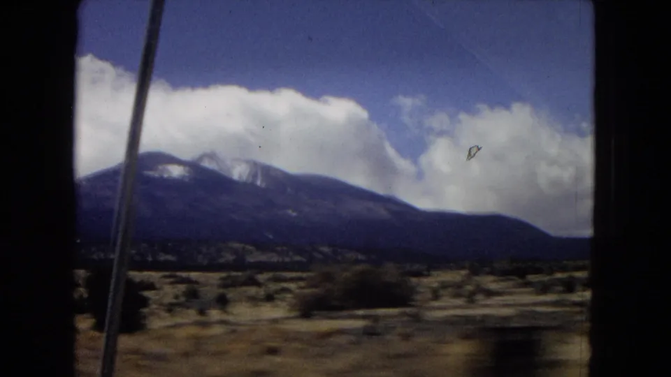 1967 home-movie frame of an arid U.S. landscape with desert scrub, distant snow-capped mountains, blue sky, white clouds, and a dark car-window edge in the foreground.