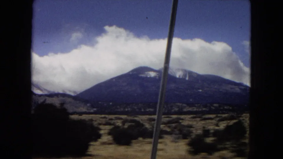 1967 home-movie frame looking from inside a vehicle toward a rounded mountain with snow patches, desert brush, bright clouds, and a slender foreground pole or antenna.