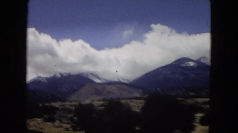 1967 home-movie frame of distant mountains partly covered by clouds, with scrubby desert vegetation and dark vehicle-window framing at the edges.