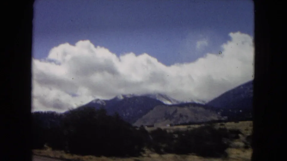 1967 home-movie frame showing desert vegetation, low hills, dark foreground framing, and distant snow-capped mountain peaks beneath large white clouds.