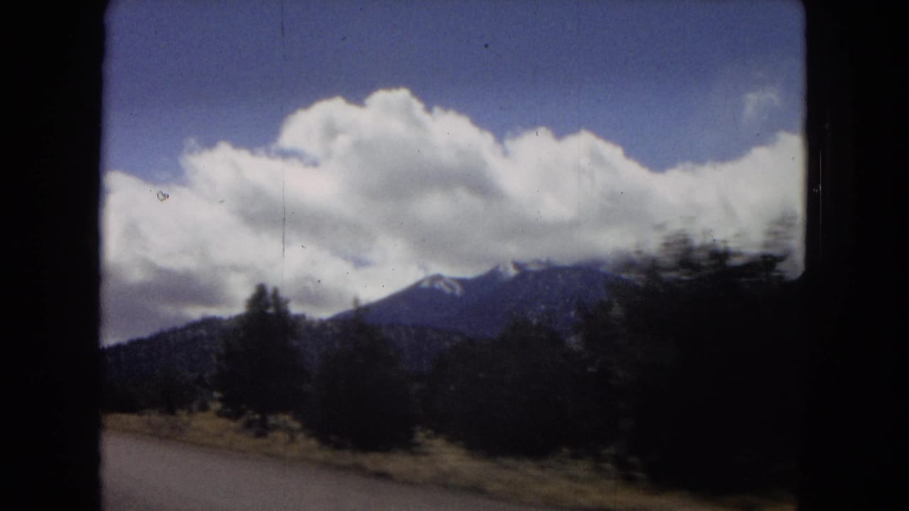 1967 Arizona home-movie frame looking from a moving car toward roadside brush, distant mountains, blue sky, and broad white clouds.