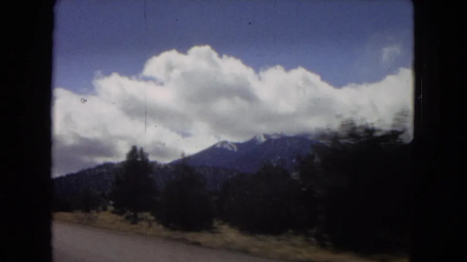 1967 Arizona home-movie frame looking from a moving car toward roadside brush, distant mountains, blue sky, and broad white clouds.